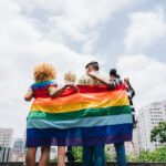 Four people stand arm in arm with their backs to the camera, holding a large rainbow Pride flag. They face a city skyline under a bright, partly cloudy sky.
