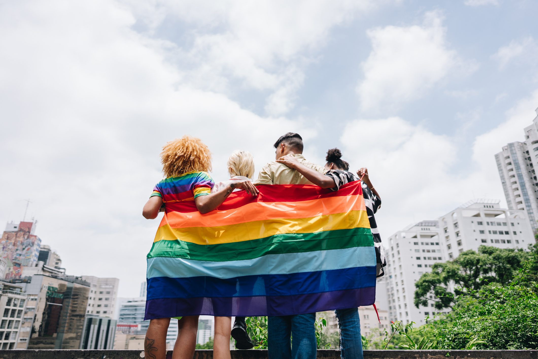 Four people stand arm in arm with their backs to the camera, holding a large rainbow Pride flag. They face a city skyline under a bright, partly cloudy sky.