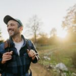 A smiling man wearing a cap and backpack walks along a trail in the countryside at sunset. He looks relaxed and happy, enjoying the outdoors.