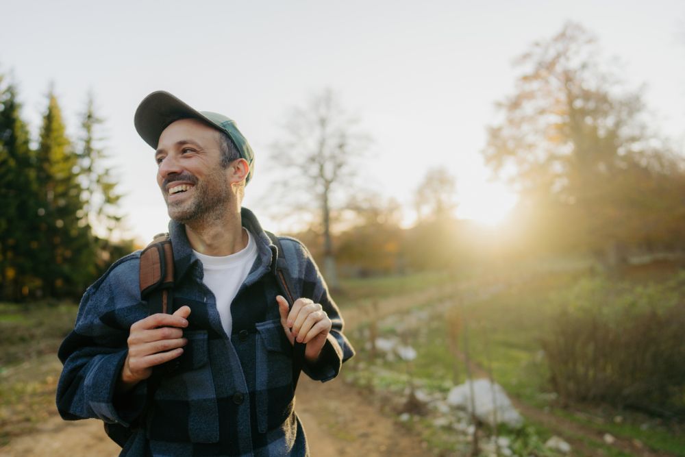 A smiling man wearing a cap and backpack walks along a trail in the countryside at sunset. He looks relaxed and happy, enjoying the outdoors.