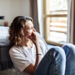 Woman sitting on the floor by a bed, looking out the window with a thoughtful, worried expression