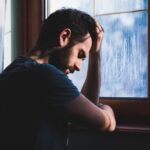 A man leans against a window with his head resting on his hand, appearing deep in thought and troubled.