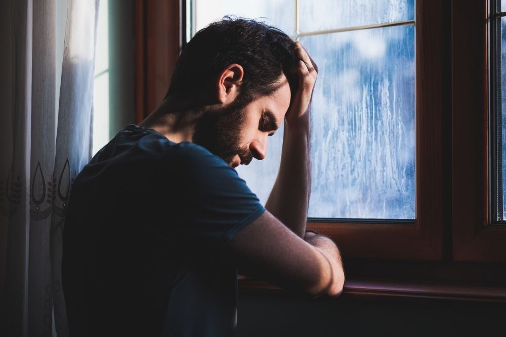 A man leans against a window with his head resting on his hand, appearing deep in thought and troubled.