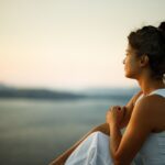 Woman sitting outdoors at sunset, looking thoughtful and calm as she gazes over the water, symbolizing reflection and mental health recovery.