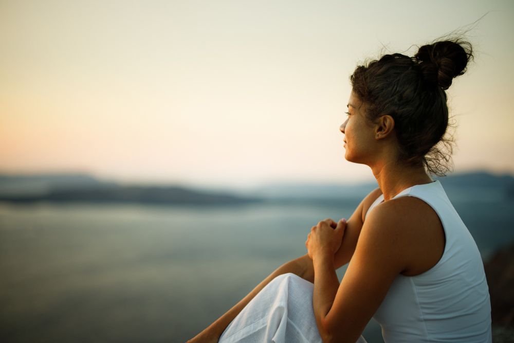 Woman sitting outdoors at sunset, looking thoughtful and calm as she gazes over the water, symbolizing reflection and mental health recovery.