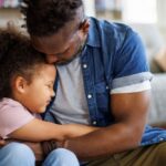 Father hugging his young daughter closely while sitting on the floor, both showing emotion and comfort.