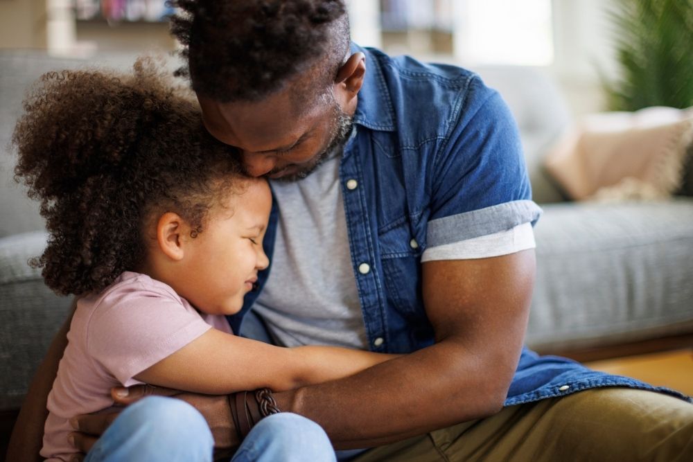 Father hugging his young daughter closely while sitting on the floor, both showing emotion and comfort.