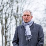 Older man wearing a gray coat and scarf standing outdoors on a cloudy winter day, looking serious, symbolizing seasonal affective disorder (SAD).