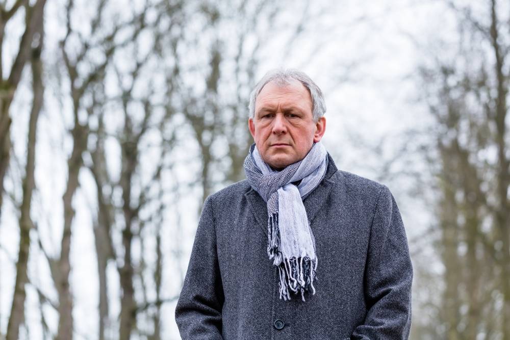 Older man wearing a gray coat and scarf standing outdoors on a cloudy winter day, looking serious, symbolizing seasonal affective disorder (SAD).