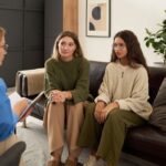 Two women sit on a couch listening attentively to a therapist who is speaking and taking notes during a counseling session.