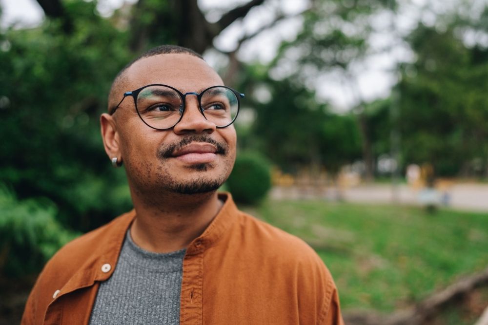 A man standing outdoors, wearing glasses and looking upward with a calm, hopeful expression.