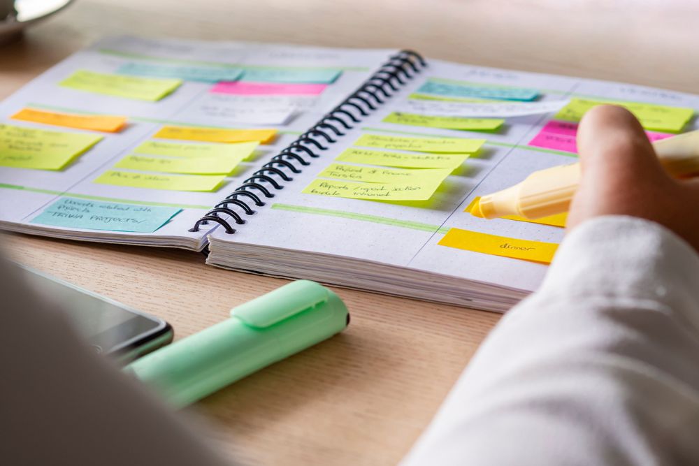 A person highlights notes in a colorful planner filled with sticky notes and schedules, symbolizing structure, routine, and organization.