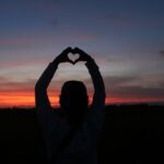 A person standing outdoors at sunset, forming a heart shape with their hands above their head, symbolizing self-love and compassion.