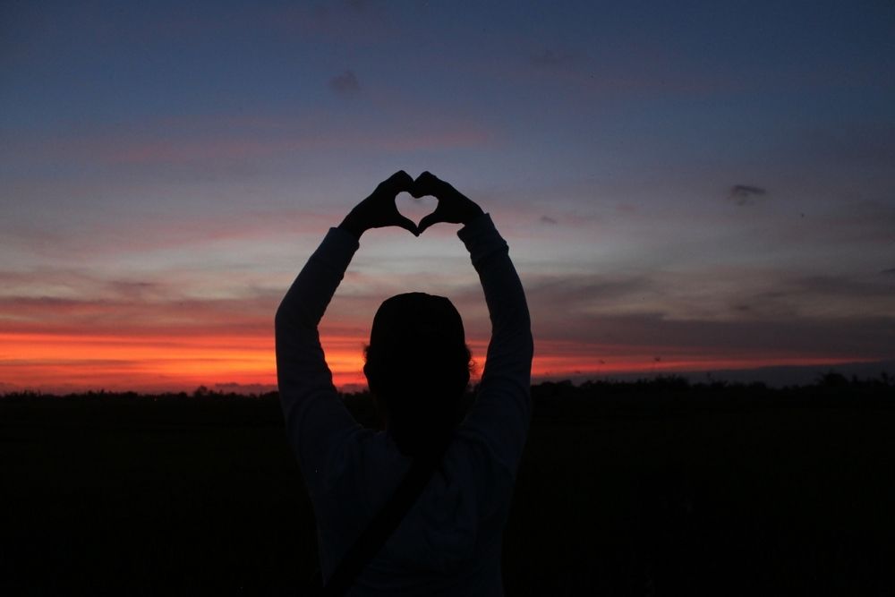 A person standing outdoors at sunset, forming a heart shape with their hands above their head, symbolizing self-love and compassion.