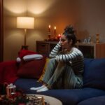 A young woman sits curled up on a couch in a dimly lit living room decorated for the holidays, looking sad and deep in thought.