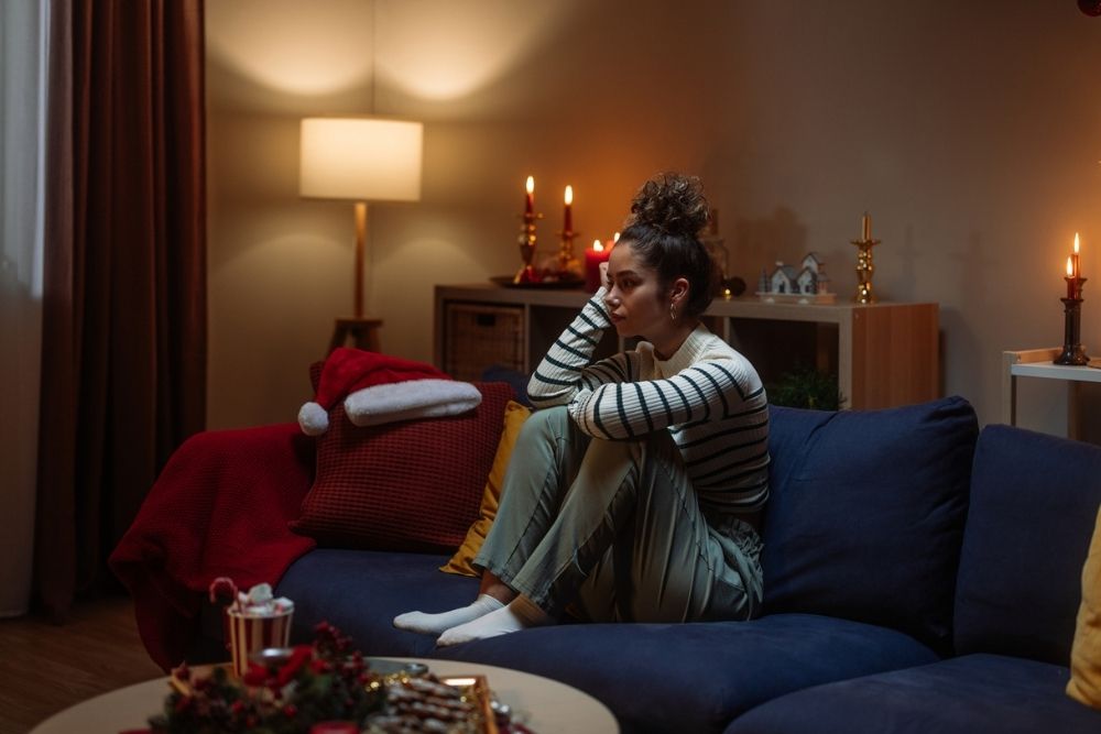 A young woman sits curled up on a couch in a dimly lit living room decorated for the holidays, looking sad and deep in thought.