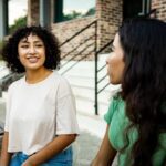 Two women sit outside on a bench, smiling and talking together, reflecting a supportive and healthy relationship in a casual setting.