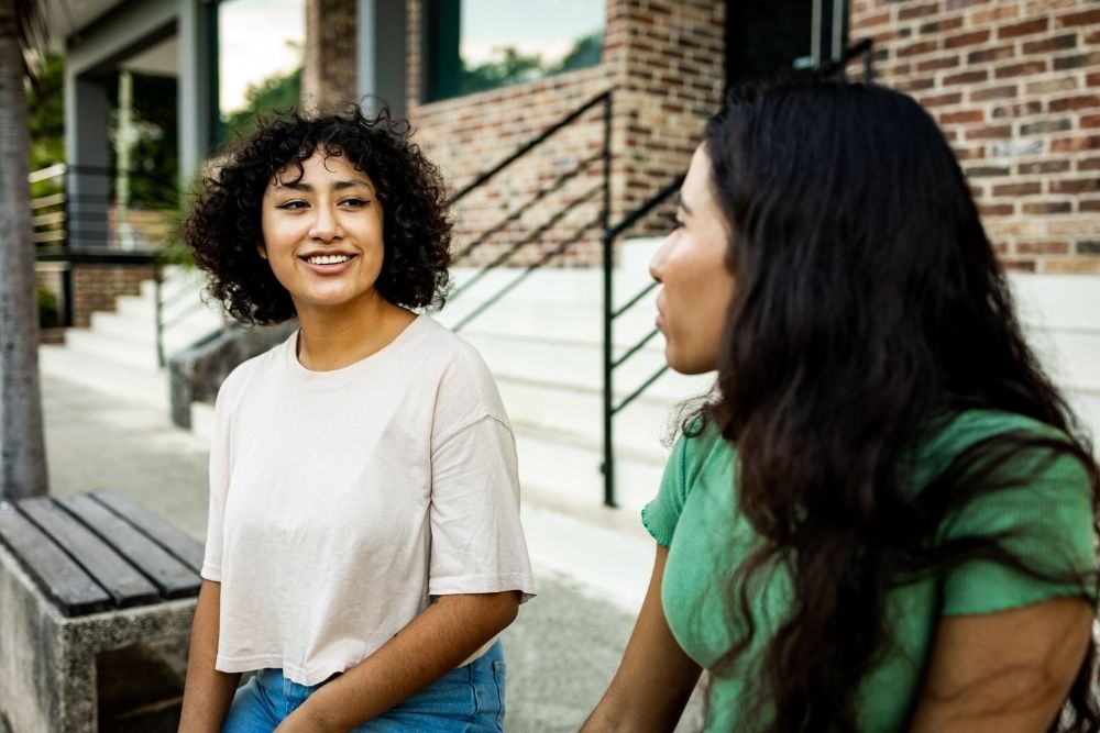 Two women sit outside on a bench, smiling and talking together, reflecting a supportive and healthy relationship in a casual setting.
