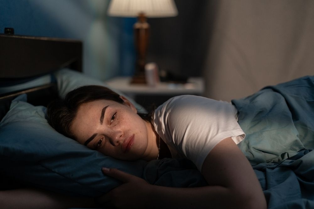 A woman lies awake in bed at night with her eyes open, appearing tired and restless in a dimly lit bedroom.
