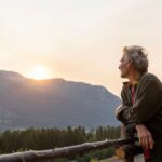 Older adult leaning on a wooden fence, watching the sun set over a mountain landscape, reflecting on hope and renewal.