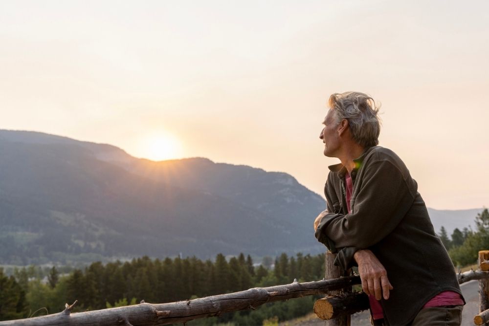 Older adult leaning on a wooden fence, watching the sun set over a mountain landscape, reflecting on hope and renewal.