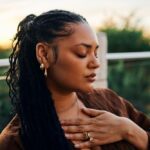 A serene woman with braided hair and golden accessories sits outdoors at sunset, her hand over her chest, exuding calm and introspection.