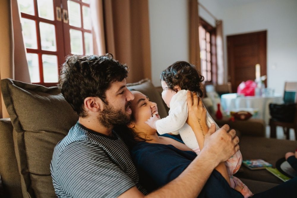 The Pavillion at Williamsburg - Tips for Creating a Supportive Home Environment After Treatment. A young family sits on a couch smiling as they hold their baby.